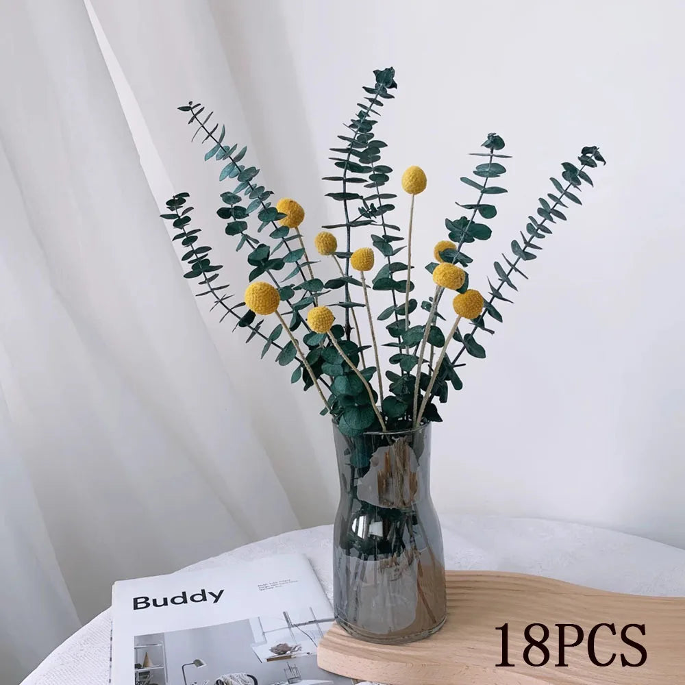 Dried eucalyptus stems with yellow billy button flowers arranged in a clear glass vase, styled on a white surface with a neutral background.
