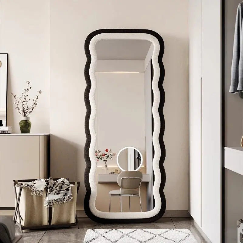 Modern full-length standing mirror with a wavy black frame, styled in a contemporary bedroom with neutral decor.
