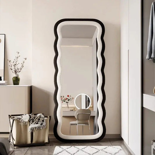 Modern full-length standing mirror with a wavy black frame, styled in a contemporary bedroom with neutral decor.
