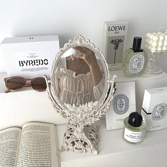 Vintage-style ornate tabletop vanity mirror with decorative frame, styled with books, sunglasses, and perfume bottles on a white surface.