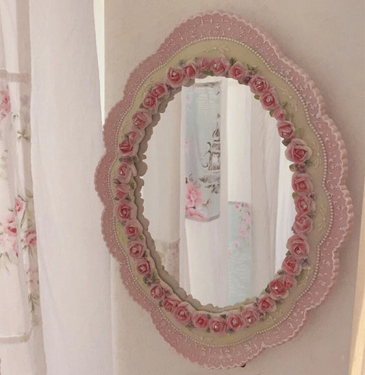 Vintage oval wall mirror with an ornate pink floral frame, featuring rose detailing and scalloped edges, styled against a soft white wall.